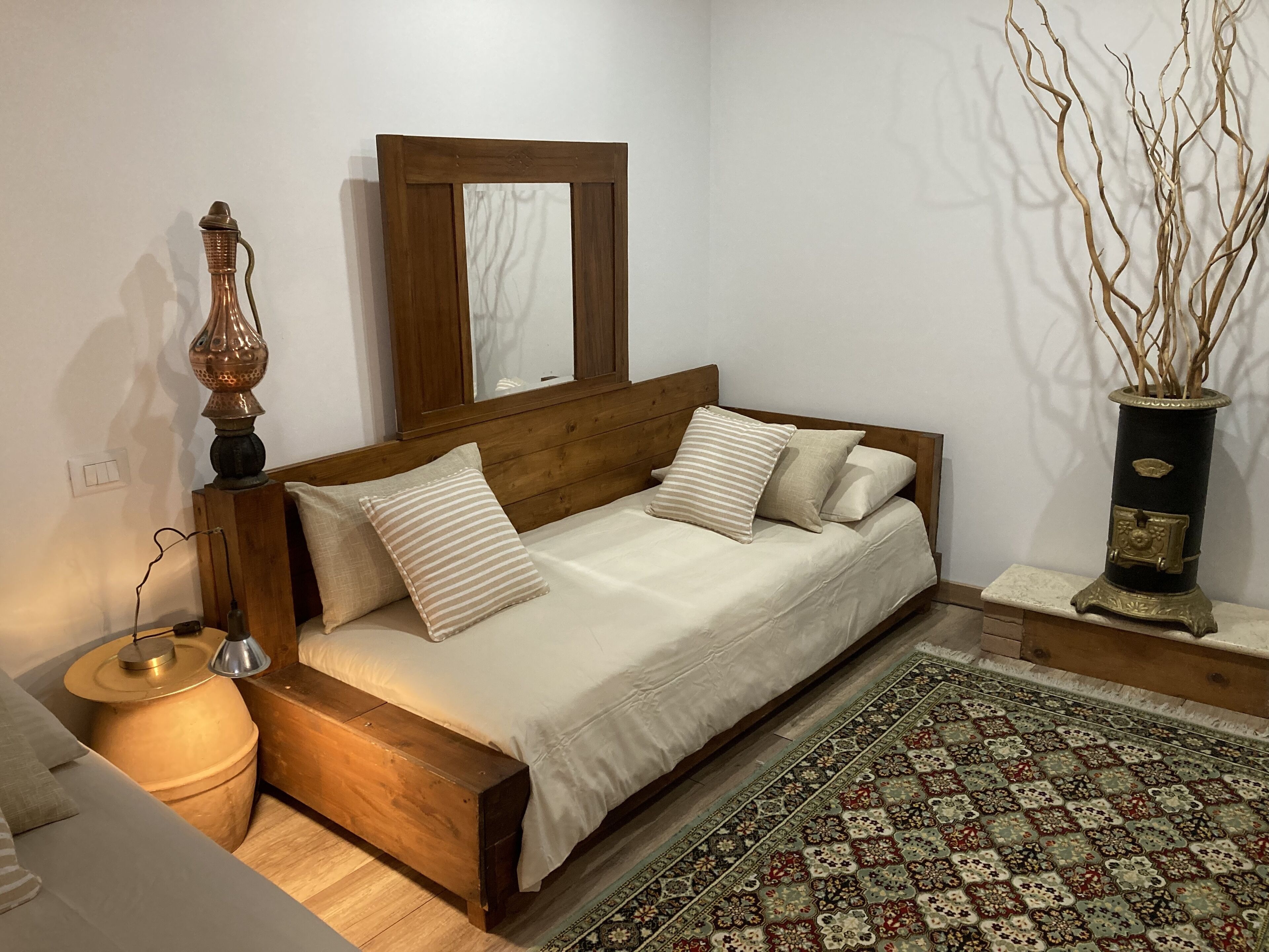 Una cama de madera con cabecera, una lámpara de noche, un florero con ramas y una alfombra estampada.