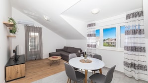 Een moderne woonkamer met een grijze bank, een eettafel met vier stoelen, een klein bijzettafeltje en een televisie op een houten kast.