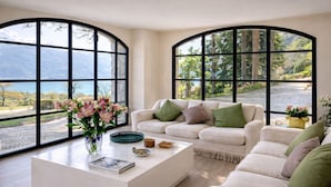 A modern living room with a white sofa set, a coffee table with decorative items, and large windows offering a view of a mountainous landscape. - Villa Gaia Luxury Retreat with Private Pool in Menaggio (Menaggio)