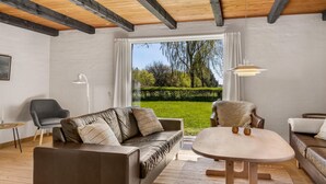 A modern living room with a brown leather sofa, a round table, and a chair. There is a large window with a view of a green garden. - 3 bedroom cozy home in Bindslev (Bindslev)