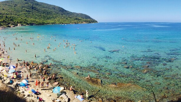 A beach with clear water, people swimming, and a mountain in the background.