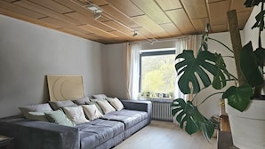A modern living room with a grey sofa, large green plant, and a window with a view of trees. - Modernes Wohnenauf dem Land (Langsur)