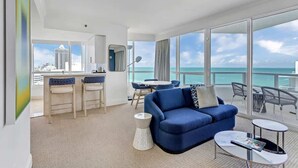 A modern hotel room with a balcony, a dining area, and a seating area with a view of the ocean and city skyline.