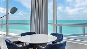 A room with a round table and four chairs, large windows with a view of the ocean, and a floor lamp.