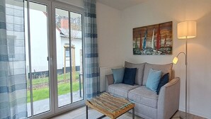A living room with a glass-top coffee table, a grey sofa with blue cushions, a floor lamp, and a painting of sailboats on the wall. - Schwedengang 7A Whg.1 Unten Links (Zingst)