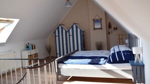 Ein Schlafzimmer mit schrägem Dach, einem Bett mit gestreifter Bettdecke, einem kleinen Nachttisch, einer Lampe und einem Fenster mit Blick nach draußen.