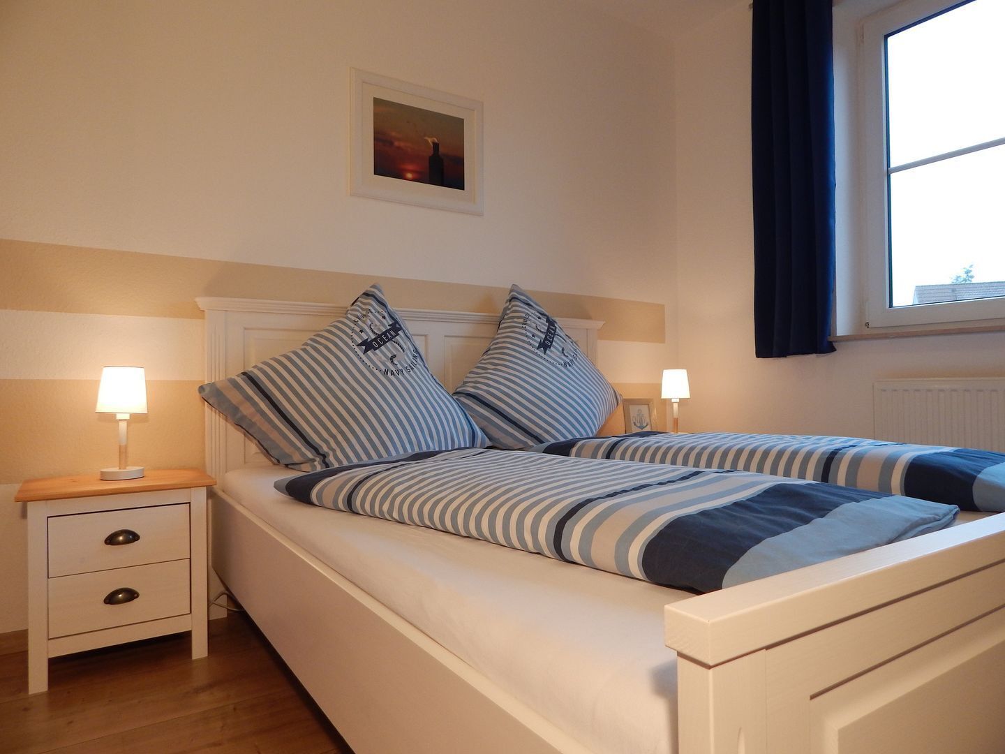 A bed with striped bedding, a bedside table with a lamp, a small drawer, and a framed picture on the wall.