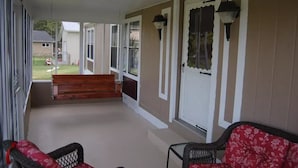 Un porche cubierto con un banco de madera, dos sillas y vista a un jardín trasero.