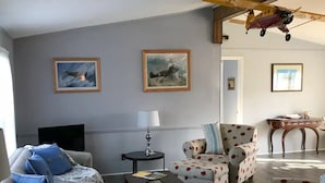 Una sala de estar con sofá, sillón y mesa de centro. Hay dos cuadros enmarcados en la pared y una mesita con una lámpara.