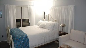 Un dormitorio con cama, mesita de noche, lámpara, una silla y ventana con cortinas.