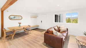 A spacious living area with a wooden dining table, a leather sofa, and a large window offering a view of the countryside. - JHRL - Tensleep A11 - Minimalist, modern slopeside condo (Teton Village)