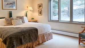 Une chambre avec un grand lit, une table de chevet, une lampe, des tableaux encadrés et une fenêtre donnant sur des arbres.