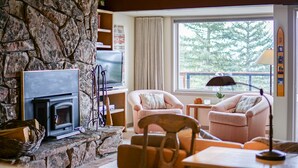 Un salon chaleureux avec une cheminée en pierre, des fauteuils confortables et une grande fenêtre donnant sur la forêt.