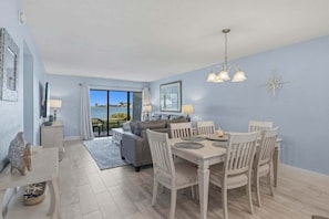 A modern living room with a sofa, dining table, and chairs, a large window with a view of the sea, and a ceiling light fixture.