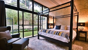 Un letto a baldacchino con vista su un giardino attraverso ampie finestre, un’area salotto con una poltrona e poggiapiedi, e un comodino con una lampada.