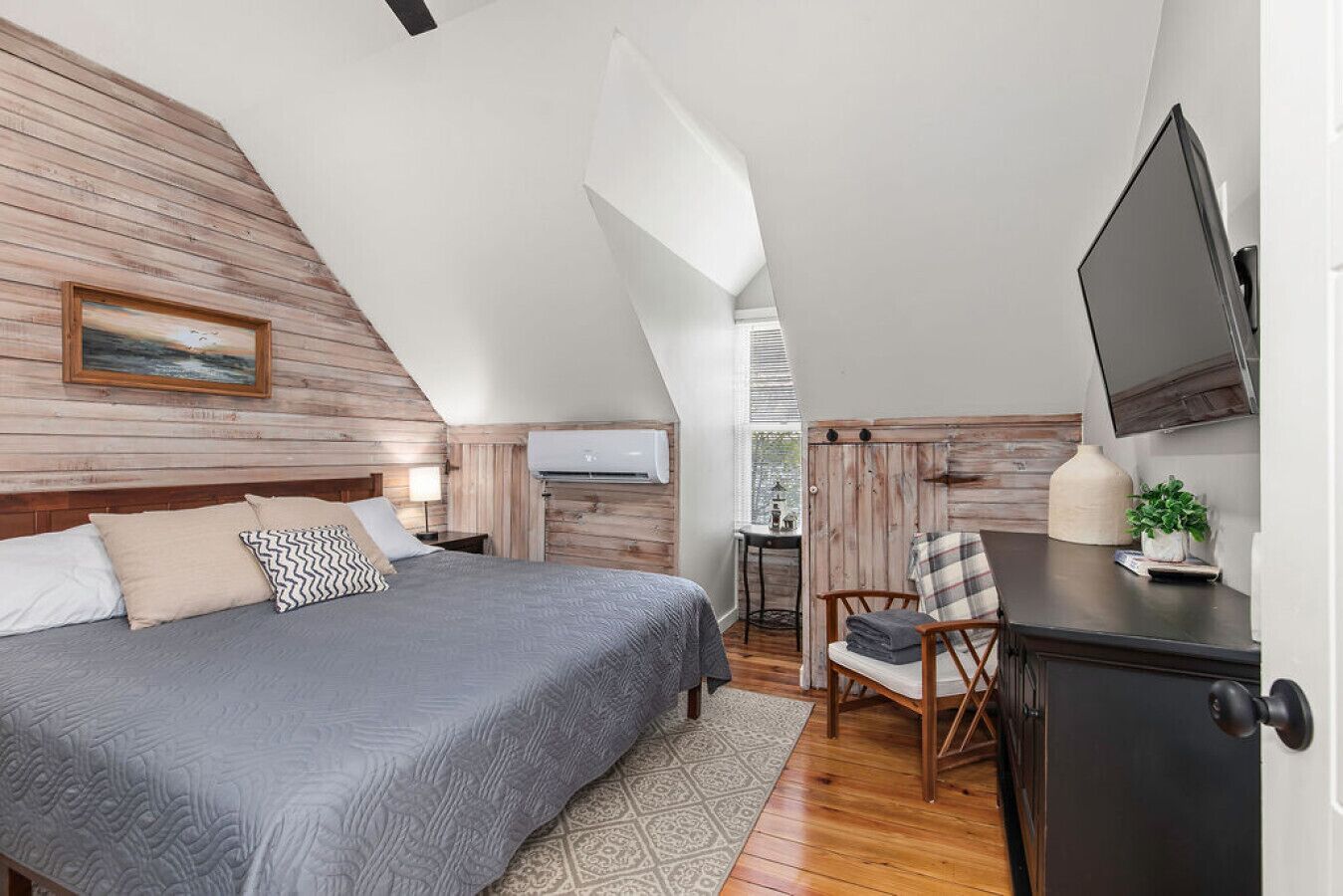 Ein Schlafzimmer mit schrägen Wänden, einem Bett mit grauer Bettdecke, einem Holz-Kopfstück, einem Schreibtisch mit Stuhl, einem Fernseher und einem Fenster mit Jalousien.