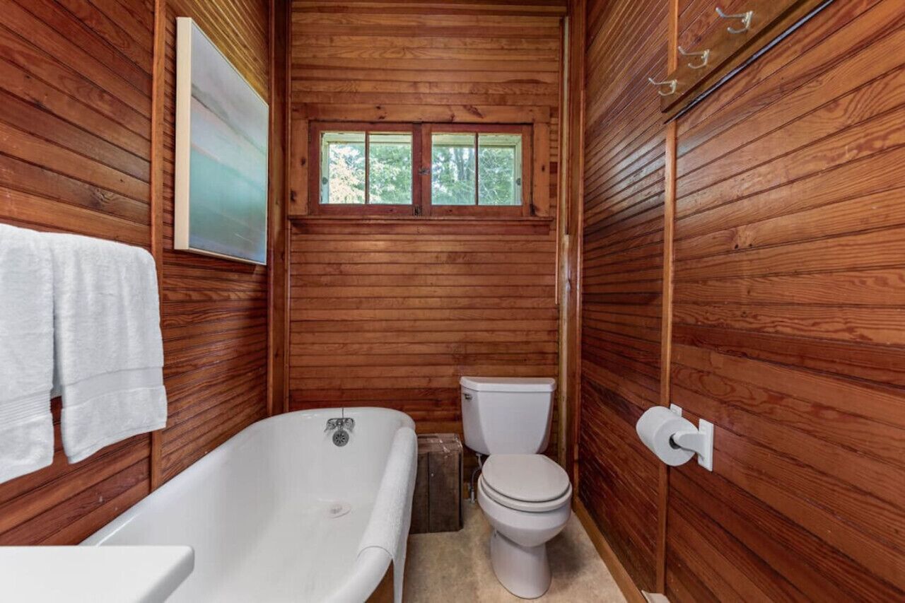 Een houten badkamer in een vakantiehuisje, met een bad, toilet en een raam.
