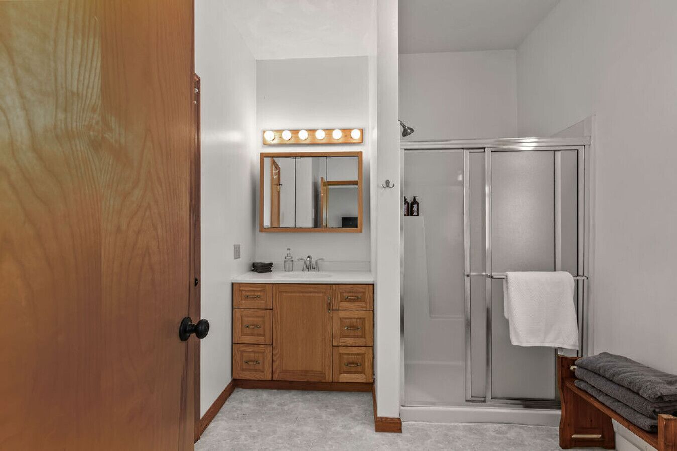 Une salle de bain avec un meuble-lavabo en bois, un miroir avec éclairage intégré, une douche et un banc avec des serviettes.