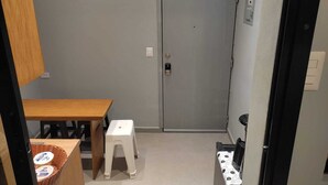 Une petite pièce avec une table en bois, un tabouret en plastique blanc, une porte et un panier contenant divers objets.