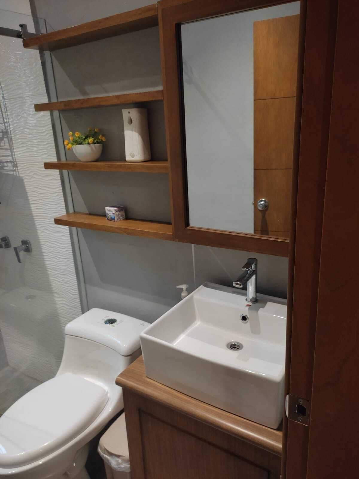 A bathroom with a toilet, a sink, and a mirror above the sink. There are wooden shelves above the sink with a bowl of flowers and a roll of toilet paper.