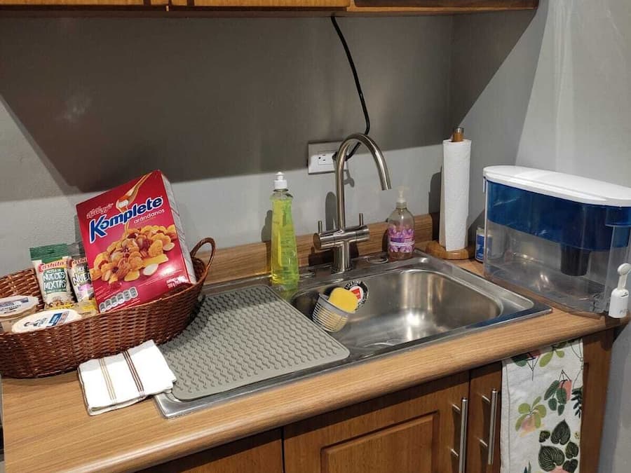A kitchen with wooden cabinets, a microwave, a sink with a soap dispenser, and a basket of snacks.