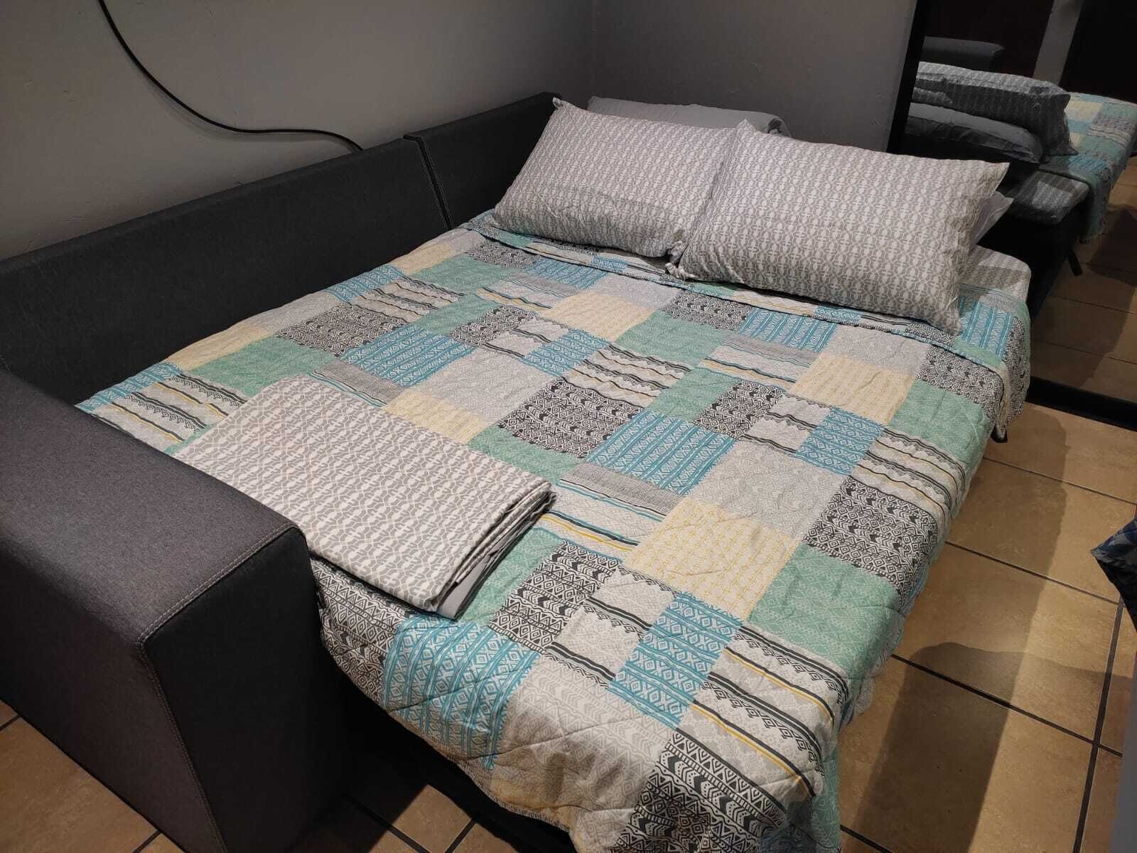 A bed with a quilted bedspread and pillows, a dark-colored sofa, and a tiled floor.