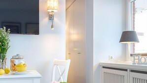 A modern kitchen with white cabinetry, a dining table with a vase of flowers, and a wall-mounted light fixture. - Strandkönig 3 - Direct beach location, mod. 1 room apartment with balcony - Andreas-Dirks (Sylt)