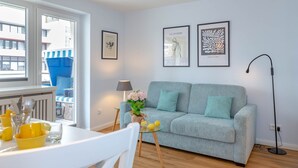 A modern living room with a grey sofa, a dining table with yellow cups, and a balcony with blue railings. - Strandkönig 3 - Direct beach location, mod. 1 room apartment with balcony - Andreas-Dirks (Sylt)