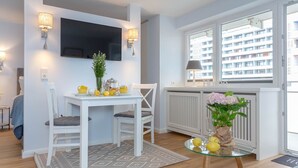 A modern hotel room with a dining area, a TV mounted on the wall, and a view of the city through large windows. - Strandkönig 3 - Direct beach location, mod. 1 room apartment with balcony - Andreas-Dirks (Sylt)