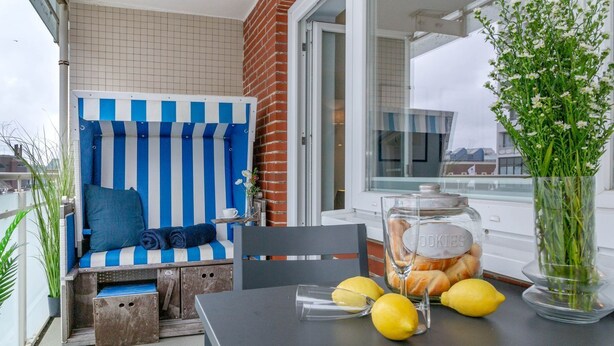 A balcony with a blue and white striped chair, a glass jar of cookies, lemons, and a vase with flowers. - Strandkönig 3 - Direct beach location, mod. 1 room apartment with balcony - Andreas-Dirks (Sylt)