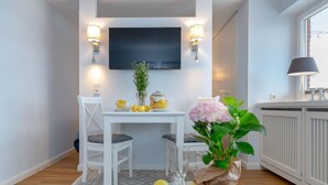 A modern kitchen with a flat-screen TV mounted above a white dining table, two chairs, and a potted plant. - Strandkönig 3 - Direct beach location, mod. 1 room apartment with balcony - Andreas-Dirks (Sylt)