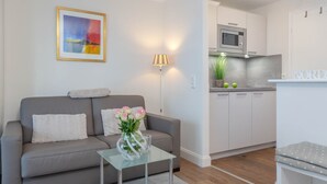 Uma sala de estar moderna com um sofá cinzento, uma mesa de centro de vidro e um vaso com flores. Um quadro está na parede, e um abajur fica ao lado do sofá. Uma cozinha com armários brancos e um micro-ondas é visível ao fundo.