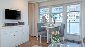 Uma sala de estar moderna com televisão de tela plana, uma mesa de jantar com cadeiras, uma pequena mesa de centro e vista da cidade através de grandes janelas.