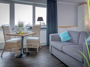 A modern living room with a grey sofa, wicker chairs, a dining table with fruits, and a view through large windows. - 5th floor, ultra-modern one-room apartment with a balcony and a view. - Andreas-Dirks (Sylt)
