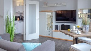 Eine moderne Küche mit integriertem Fernseher, ein Essbereich mit Tisch und Stühlen sowie ein graues Sofa im Wohnzimmer.