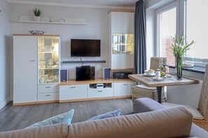 A modern living room with a flat-screen TV, built-in shelving, a dining area with a table, and a large window. - 5th floor, ultra-modern one-room apartment with a balcony and a view. - Andreas-Dirks (Sylt)
