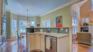A kitchen with a central island, wooden cabinets, a dining area with chairs, and a view of the outdoors. - Ingen Trafikk secluded forest oasis (Bald Head Island)