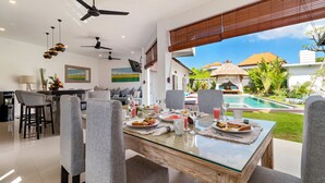 A modern dining area with a glass-top table, grey upholstered chairs, ceiling fans, and a view of a swimming pool and tropical garden. - Luxurious 4BR Villa with Pool, Bali Villa 2206 (Bali)