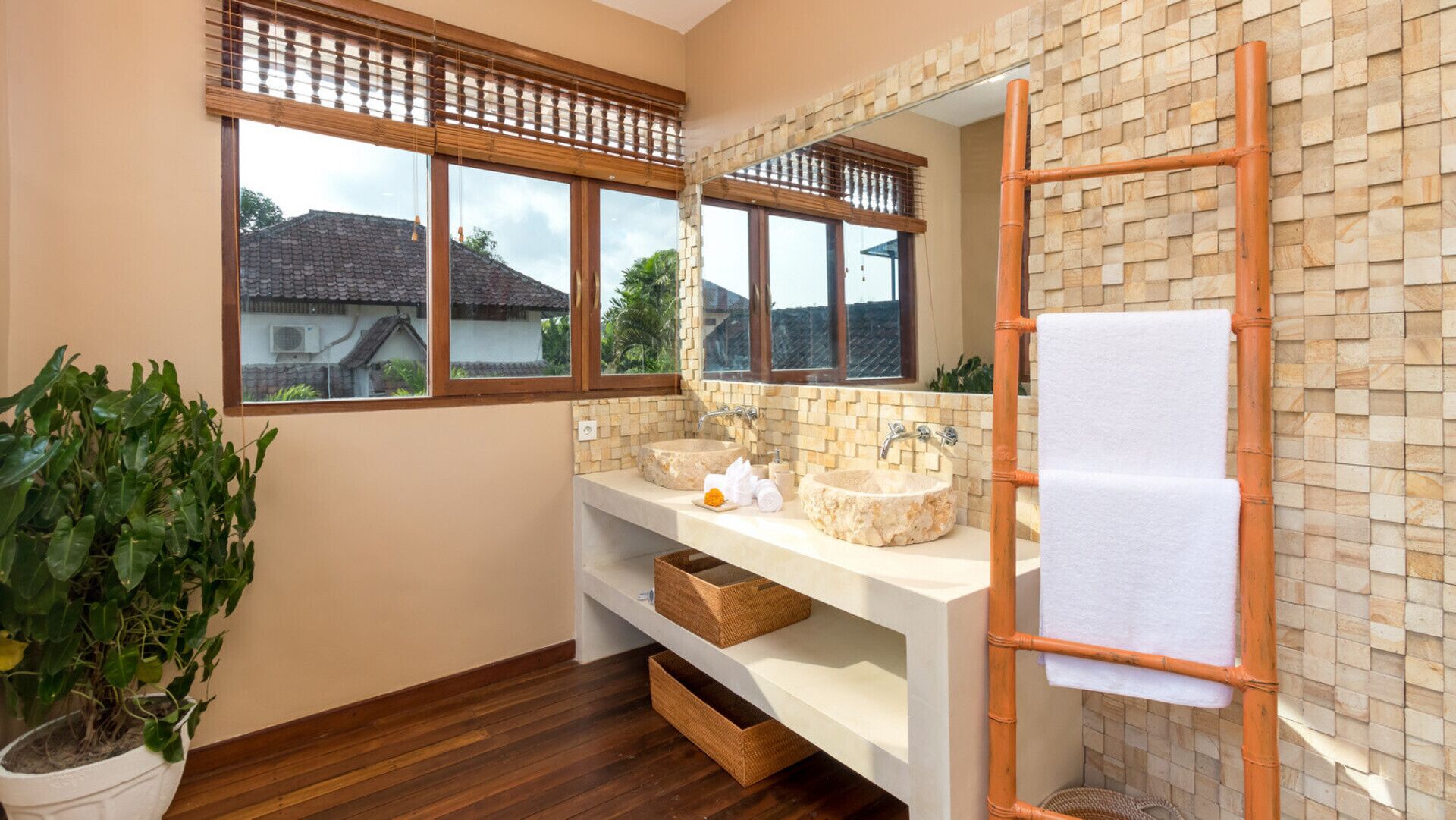 A bathroom with two stone sinks, a wooden ladder, a large mirror, and a view of houses through the windows.