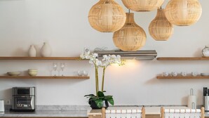 Et køkken med et moderne design, med indbygget ovn, kaffemaskine og en træbordplade. Der hænger fletvæglamper over bordet, og der står en orkidé i en potte på bordpladen.