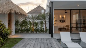Ett modernt hus med en träterrass, en byggnad med halmtak och ett inglasat vardagsrum med en soffa och fåtöljer.