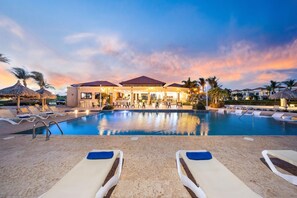 Zona de piscina en un resort con sillones, un bar y un edificio de techo rojo.