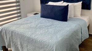 A neatly made bed with a blue headboard and white bedding, a bedside table with a remote control, and a window with blinds. - Departamento con alberca en El Encanto (Mazatlán)