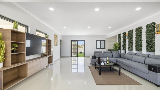 A spacious living room with a large grey sofa, a flat-screen TV on a wooden stand, and a coffee table. - *NEW* 4BR4BA Villa across of PalmBeach w/ Pvt Pool (Noord)