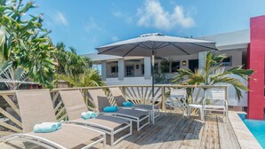 A poolside area with lounge chairs and an umbrella, surrounded by palm trees and a building with a red wall. - Quiet, Cozy & Central ~ Close to the Beach ~ Pool! (Noord)