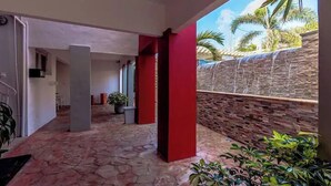 A covered outdoor area with stone flooring, red pillars, and a view of a stone wall and palm trees. - Quiet, Cozy & Central ~ Close to the Beach ~ Pool! (Noord)