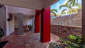 A covered outdoor area with a stone floor, red pillars, and a view of palm trees and a stone wall. - 2BR 1BA Ground Level in the Heart of Aruba ~ Pool (Noord)