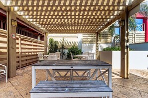 Ulkoterassi, jossa on puinen pöytä ja tuolit, yläpuolella oleva pergola ja taustalla näkyvä rakennus.
