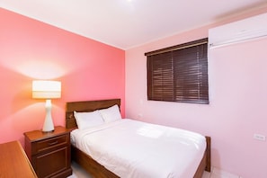 A bedroom with a bed, a nightstand, a lamp, a window with blinds, and an air conditioning unit. - 2BR 1BA Ground Level in the Heart of Aruba ~ Pool (Noord)