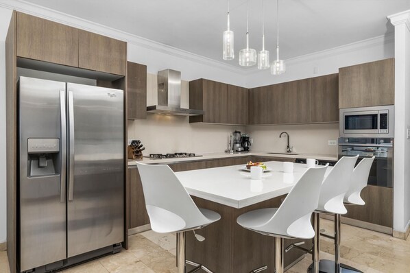 Cocina moderna con refrigerador de acero inoxidable, horno empotrado, microondas y cafetera. Hay tres taburetes blancos alrededor de una isla central.